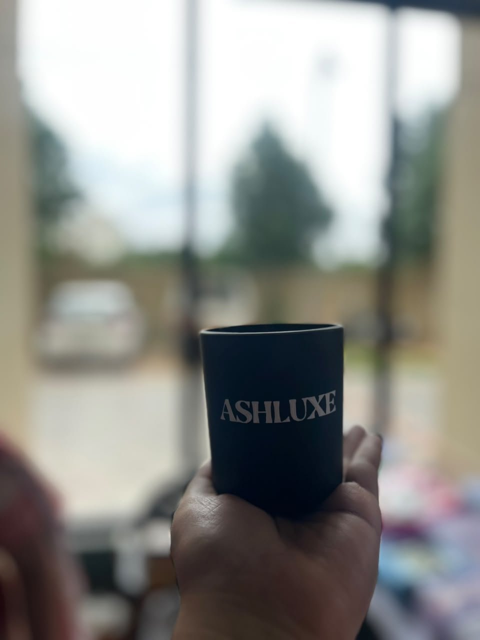Hand holding a matte black ASHLUXE candle by a window